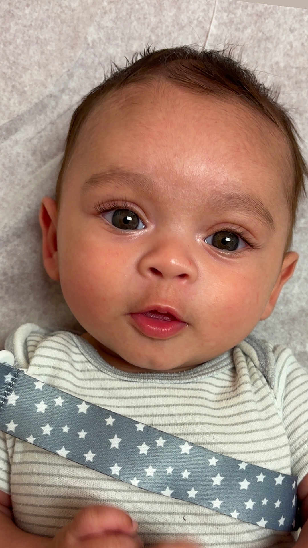 Yadiel is registered to the contest to win money with this photo: baby, infant, face, eyes, onesie, striped_clothing, star_pattern, strap, skin, head, cheeks, mouth, expression, lying_down, closeup, cute, young_child, portrait, person, indoors