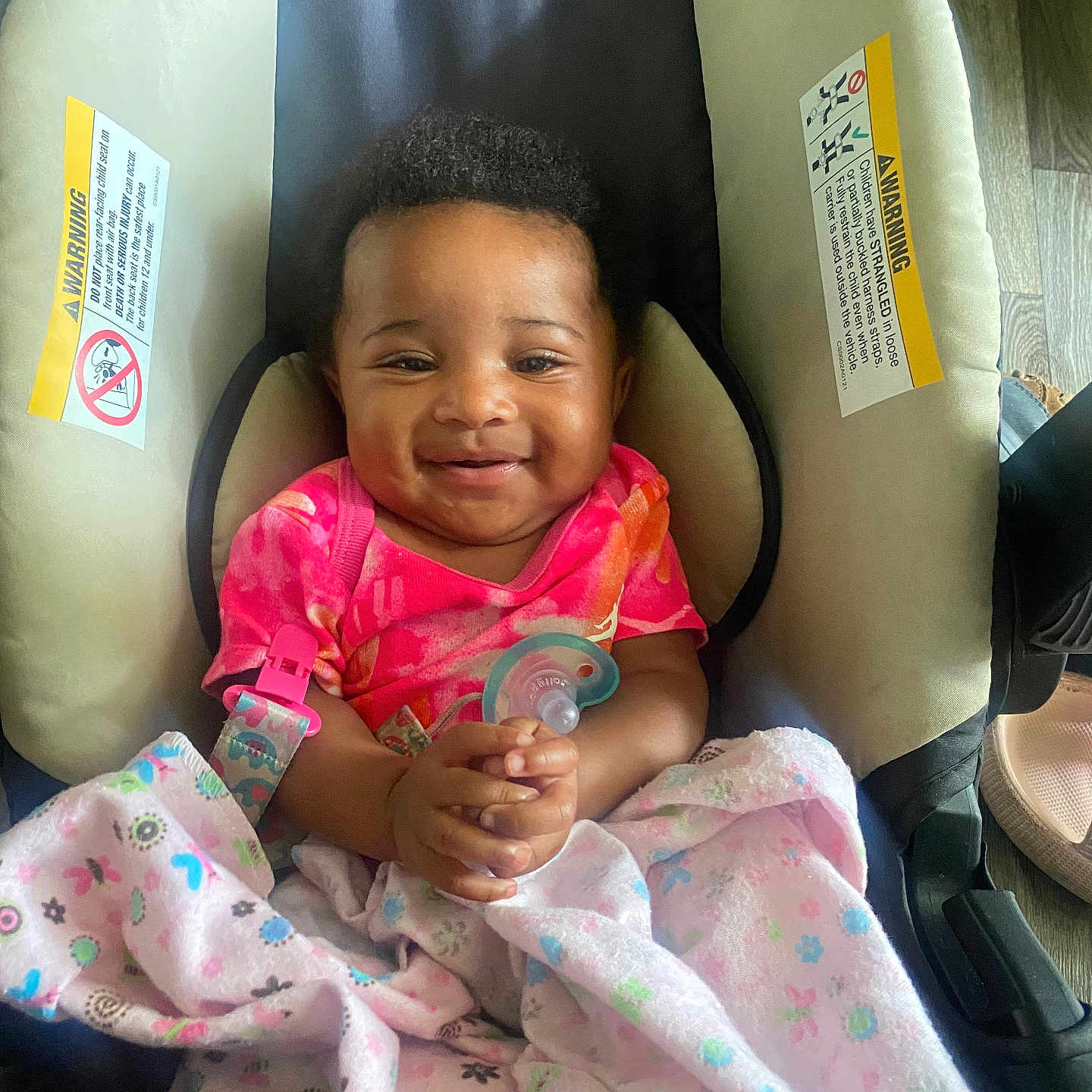 Amor is registered to the contest to win money with this photo: baby, smiling, car_seat, pacifier, blanket, pink_clothing, infant, child, happy, indoor, floor, wooden_floor, warning_label, cushion, seat, person, toddler, cute, holding, wrapped