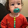 Carlos is registered to the contest to win money with this photo: toddler, child, pacifier, orange_sweater, beads, indoor, curly_hair, snack_bowl, hand, face, expression, sitting, cute, young_child, casual_clothing, portrait, curious, home, person, baby