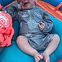 baby, sleeping, smiling, blue_cushion, orange_mesh, grey_outfit, infant, relaxed, hand_on_face, barefoot, cute, patterned_clothing, resting, child, portrait, indoors, soft_lighting, peaceful, small_person, comfort