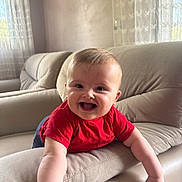Enes is registered to the contest to win money with this photo: baby, child, smiling, red_shirt, couch, living_room, arm, happy, indoor, furniture, curtains, natural_light, infant, face, person, portrait, home, cute, joy, leaning