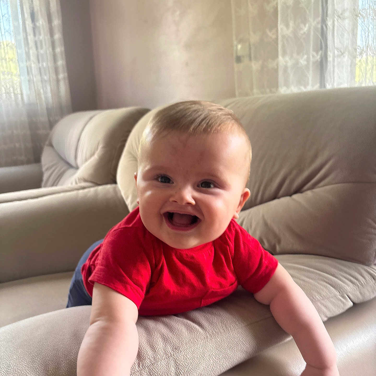 Enes is registered to the contest to win money with this photo: arm, baby, child, couch, curtains, cute, face, furniture, happy, home, indoor, infant, joy, leaning, living_room, natural_light, person, portrait, red_shirt, smiling