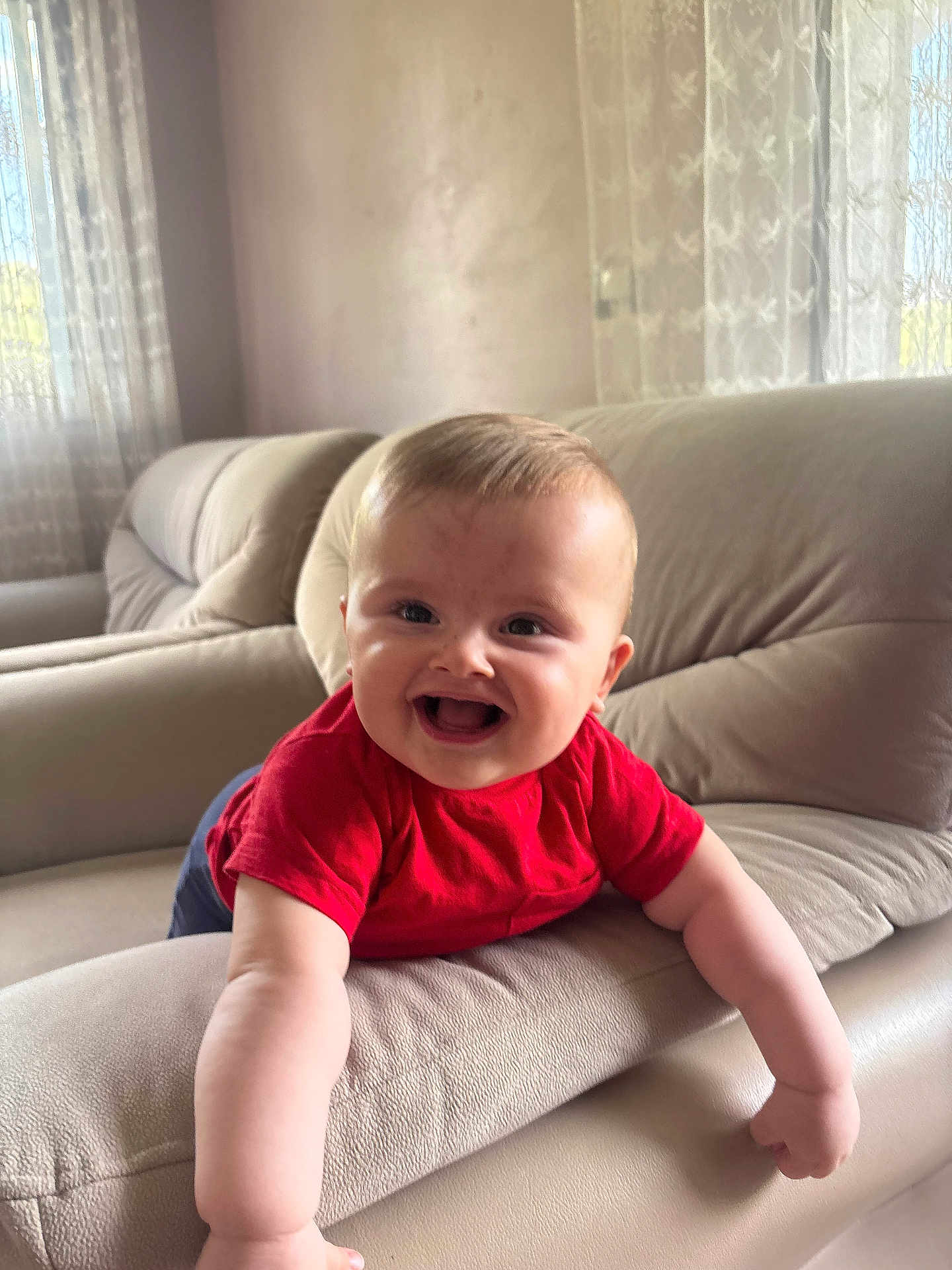 Enes is registered to the contest to win money with this photo: baby, child, smiling, red_shirt, couch, living_room, arm, happy, indoor, furniture, curtains, natural_light, infant, face, person, portrait, home, cute, joy, leaning