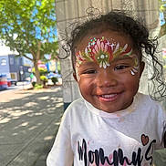 Monaya joined the competition — help win amazing prizes! child, toddler, face_paint, smiling, curly_hair, outdoor, sunlight, sidewalk, white_shirt, text_on_clothing, happy, portrait, urban, tree, daylight, person, cute, young_child, colorful, casual_wear