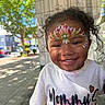 child, toddler, face_paint, smiling, curly_hair, outdoor, sunlight, sidewalk, white_shirt, text_on_clothing, happy, portrait, urban, tree, daylight, person, cute, young_child, colorful, casual_wear