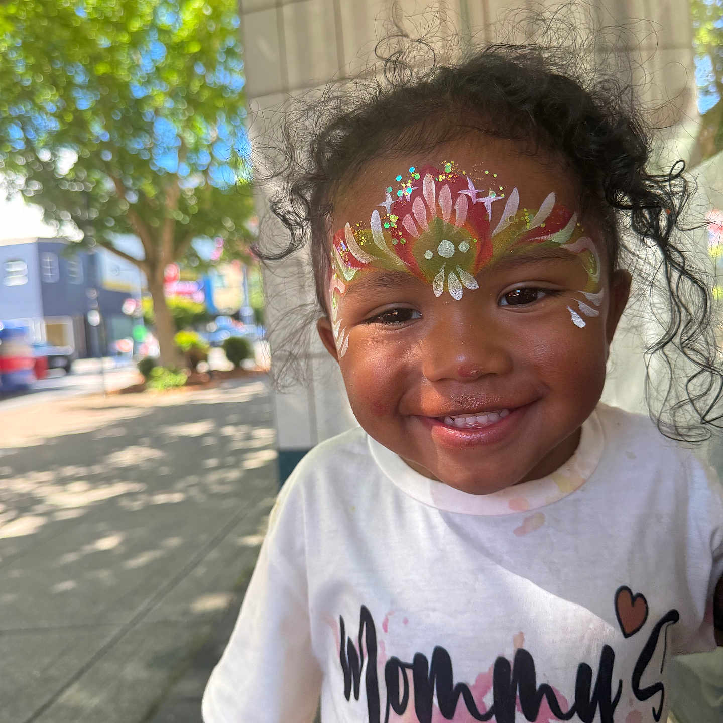 Monaya joined the competition — help win amazing prizes! child, toddler, face_paint, smiling, curly_hair, outdoor, sunlight, sidewalk, white_shirt, text_on_clothing, happy, portrait, urban, tree, daylight, person, cute, young_child, colorful, casual_wear