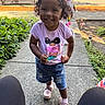 cars, casual_clothing, child, curly_hair, daytime, denim_shorts, greenery, happy, outdoor, person, pink_shirt, plants, play, shoes, sidewalk, smiling, suburban, sunlight, toddler, tricycle