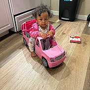 Monaya is registered to the contest to win money with this photo: book, cabinet, child, curious, curly_hair, floor_mat, footwearless, indoor, kitchen, person, pink, play, serious_expression, small_vehicle, stove, toddler, toy_car, trash_bin, wooden_floor, young_child