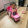 book, cabinet, child, curious, curly_hair, floor_mat, footwearless, indoor, kitchen, person, pink, play, serious_expression, small_vehicle, stove, toddler, toy_car, trash_bin, wooden_floor, young_child