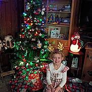 Allison is registered to the contest to win money with this photo: child, christmas_tree, presents, holiday_decorations, reindeer_antlers, plaid_dress, smile, living_room, festive_lights, ornaments, carpet, wooden_furniture, snowman_decoration, cabinet, television, wrapped_gifts, christmas_lights, holiday_spirit, indoor, happy