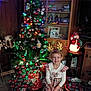 child, christmas_tree, presents, holiday_decorations, reindeer_antlers, plaid_dress, smile, living_room, festive_lights, ornaments, carpet, wooden_furniture, snowman_decoration, cabinet, television, wrapped_gifts, christmas_lights, holiday_spirit, indoor, happy