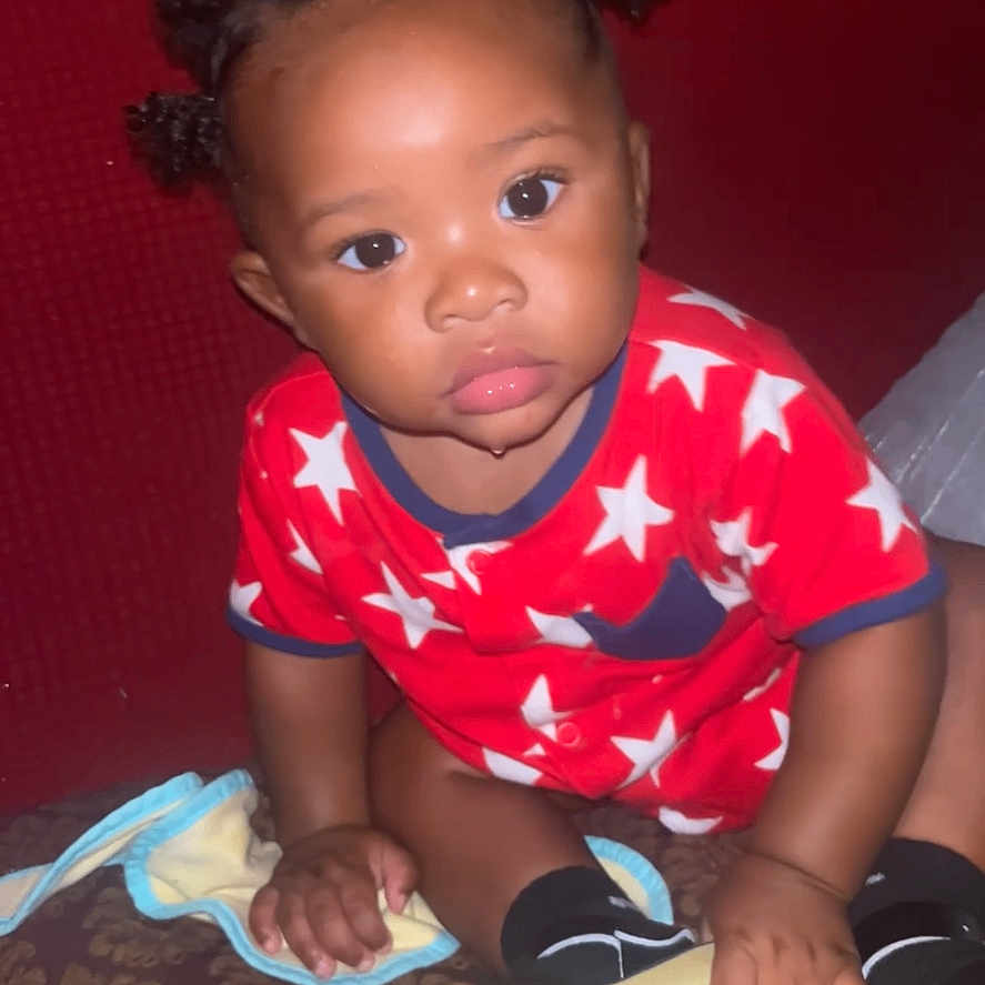 Kaden joined the competition — help win amazing prizes! toddler, child, baby, pajamas, stars, hair_buns, sandal, indoor, curious, patterned_surface, hand, footwear, person, cute, young_child, sitting, looking, bright_eyes, red_clothing, black_socks