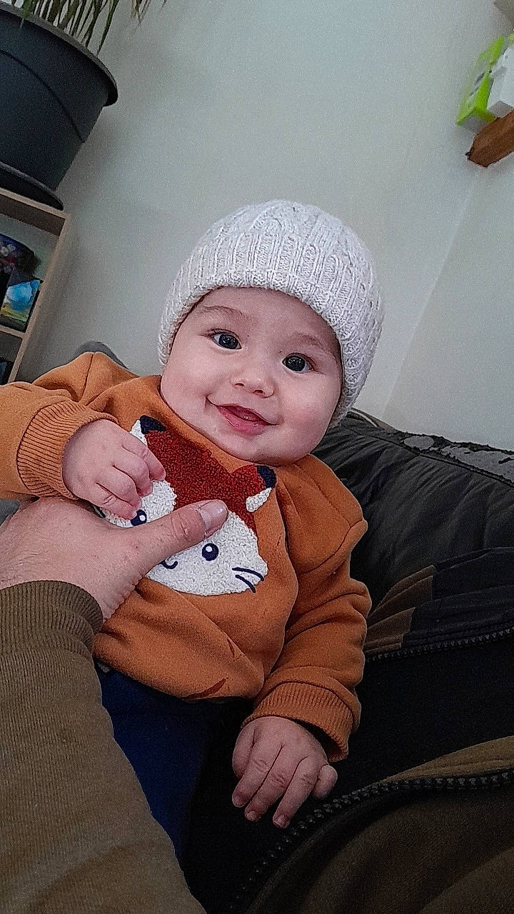 Eros participe au concours pour gagner de l'argent avec cette photo : baby, baby_toddler_clothing, beanie, cap, cheek, child, comfort, eye, fashion_accessory, fun, gesture, happy, headwear, joy, knit_cap, lap, person, sitting, skin, sleeve