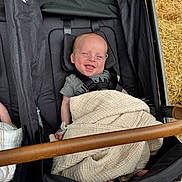 Greyson is registered to the contest to win money with this photo: baby, blanket, stroller, smiling, child, infant, outdoor, seatbelt, black_clothing, head_support, brown_bar, fabric, person, happy, cute, small, toddler, comfort, daylight, relaxing