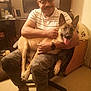 bald, camo_pants, casual_clothing, chair, computer_monitor, desk, dog, floor, furniture, glasses, happy, indoor, man, pet, pet_bed, room, slippers, smiling, warm_lighting, watch