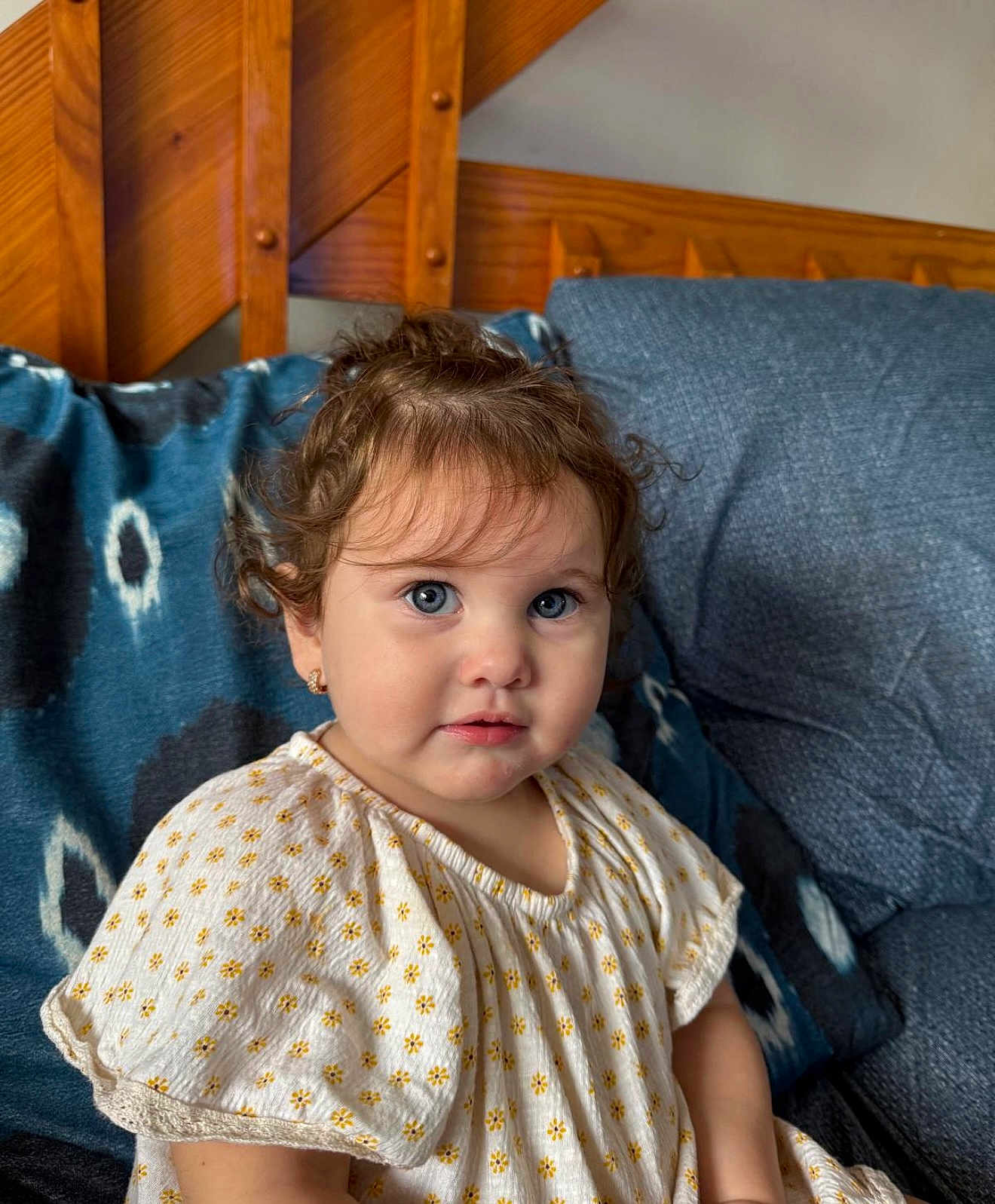 Hannah joined the competition — help win amazing prizes! child, toddler, curly_hair, blue_eyes, dress, patterned_clothing, couch, cushion, wooden_background, indoor, portrait, cute, sitting, young_child, person, face, smiling, earrings, home, cozy