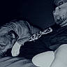 animal, beard, bond, cap, casual, closeup, comfort, dark_background, dog, friendship, home, indoor, laying_down, man, monochrome, pet, portrait, relaxation, resting, shirt