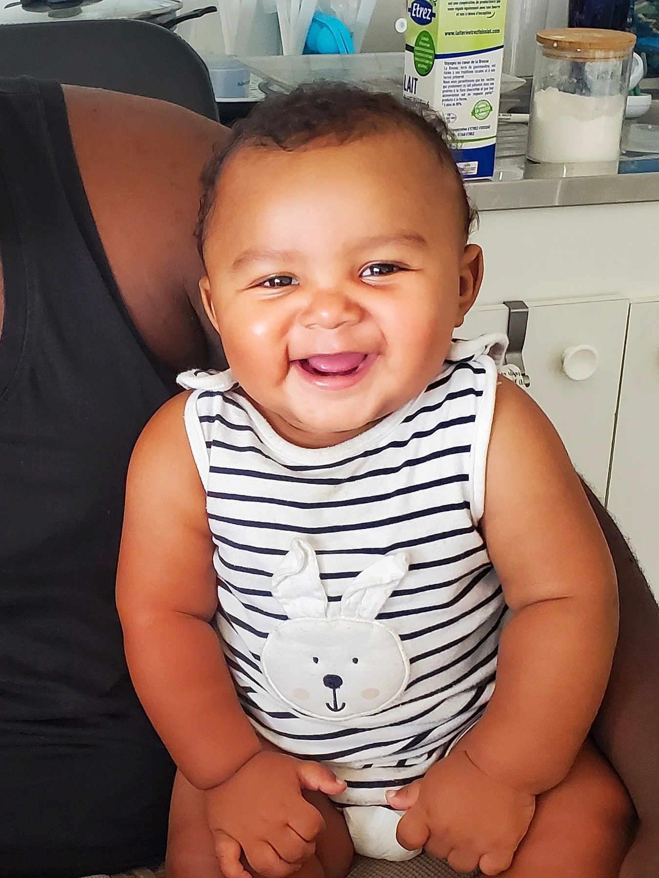 Lovan participe au concours pour gagner de l'argent avec cette photo : baby, smiling, striped_shirt, bunny_design, sitting, person, arm, kitchen, milk_carton, container, cabinet, furniture, happy, child, indoor, cute, portrait, infant, hand, face