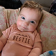 Isla is registered to the contest to win money with this photo: baby, infant, smile, blue_eyes, short_sleeve_shirt, text_on_clothing, blanket, floral_pattern, soft_texture, indoor, closeup, chubby_cheeks, cute, person, portrait, hand, toy, cozy, relaxed, happy