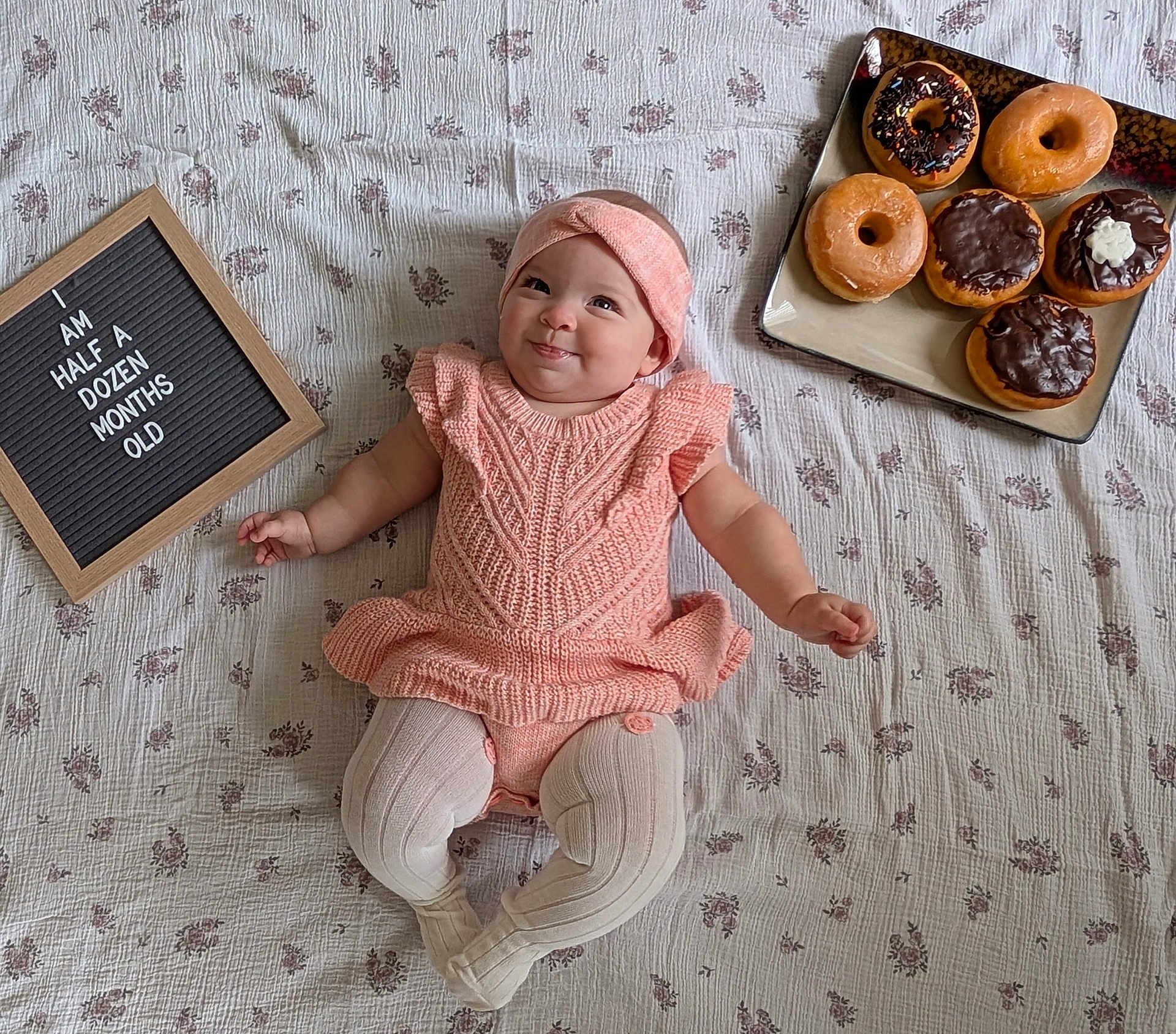 Emilia joined the competition — help win amazing prizes! baby, infant, smile, headband, knitwear, blanket, floral_pattern, sign, text, donuts, plate, food, milestone, candid, cute, child, laying_down, indoors, celebration, person