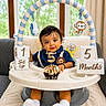baby, infant, milestone_blocks, cupcake, number_candle, baby_seat, toy, hanging_toys, blue_outfit, white_socks, indoors, window, curtains, couch, blanket, smiling, portrait, child, celebration, cute