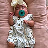 baby, infant, pacifier, headband, bow, floral_dress, pink_cushion, sleeping, medical_sensor, ankle_band, soft_texture, cute, resting, indoors, child, newborn, comfort, peaceful, small_hands, tiny_feet