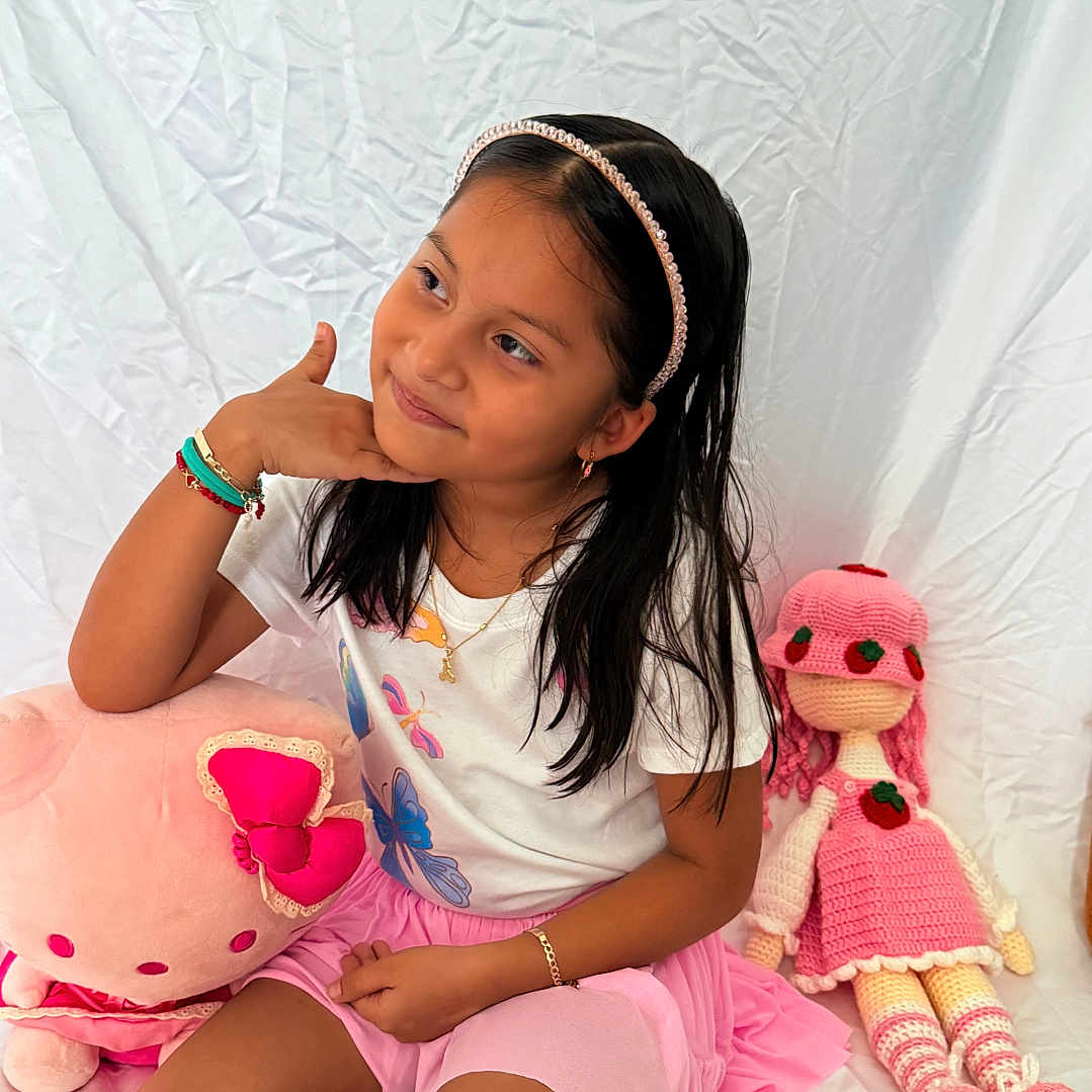 Yeimy is registered to the contest to win money with this photo: child, girl, portrait, sitting, smiling, indoors, bed, pink, doll, stuffed_animal, plush_toy, headband, long_hair, bracelets, necklace, skirt, toy, white_background, fashion, playroom