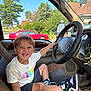 Kayson is registered to the contest to win money with this photo: child, smiling, car_interior, steering_wheel, seat, window, tshirt, shorts, sneakers, happy, boy, seatbelt, dashboard, trees, red_car, daylight, playful, vehicle, inside_car, casual_clothing