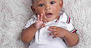 Keanu is registered to the contest to win money with this photo: baby, child, curly_hair, white_polo_shirt, blue_shorts, fluffy_blanket, hand_gesture, cute, portrait, infant, smiling, indoors, happy, lying_down, skin, face, eyes, mouth, clothing, young