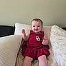 Hazel is registered to the contest to win money with this photo: baby, child, smiling, dress, red_dress, sitting, pillow, couch, indoor, happy, person, infant, cute, furniture, face, head, skin, feet, hand, young
