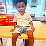 Kynd joined the competition — help win amazing prizes! child, toddler, boy, afro_hair, white_tshirt, denim_shorts, sneakers, table, indoor, window, blurred_background, casual_pose, colorful_sign, light, floor, seated, looking_at_camera, young, cute, fashion