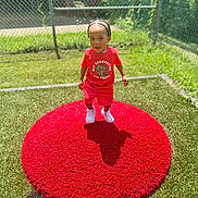 Kynd is registered to the contest to win money with this photo: toddler, child, red_clothing, white_sneakers, red_mat, outdoor, grass, sunny, fence, playground, happy, walking, young_child, summer, daylight, person, shorts, tshirt, braided_hair, shadow