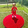 toddler, child, red_clothing, white_sneakers, red_mat, outdoor, grass, sunny, fence, playground, happy, walking, young_child, summer, daylight, person, shorts, tshirt, braided_hair, shadow