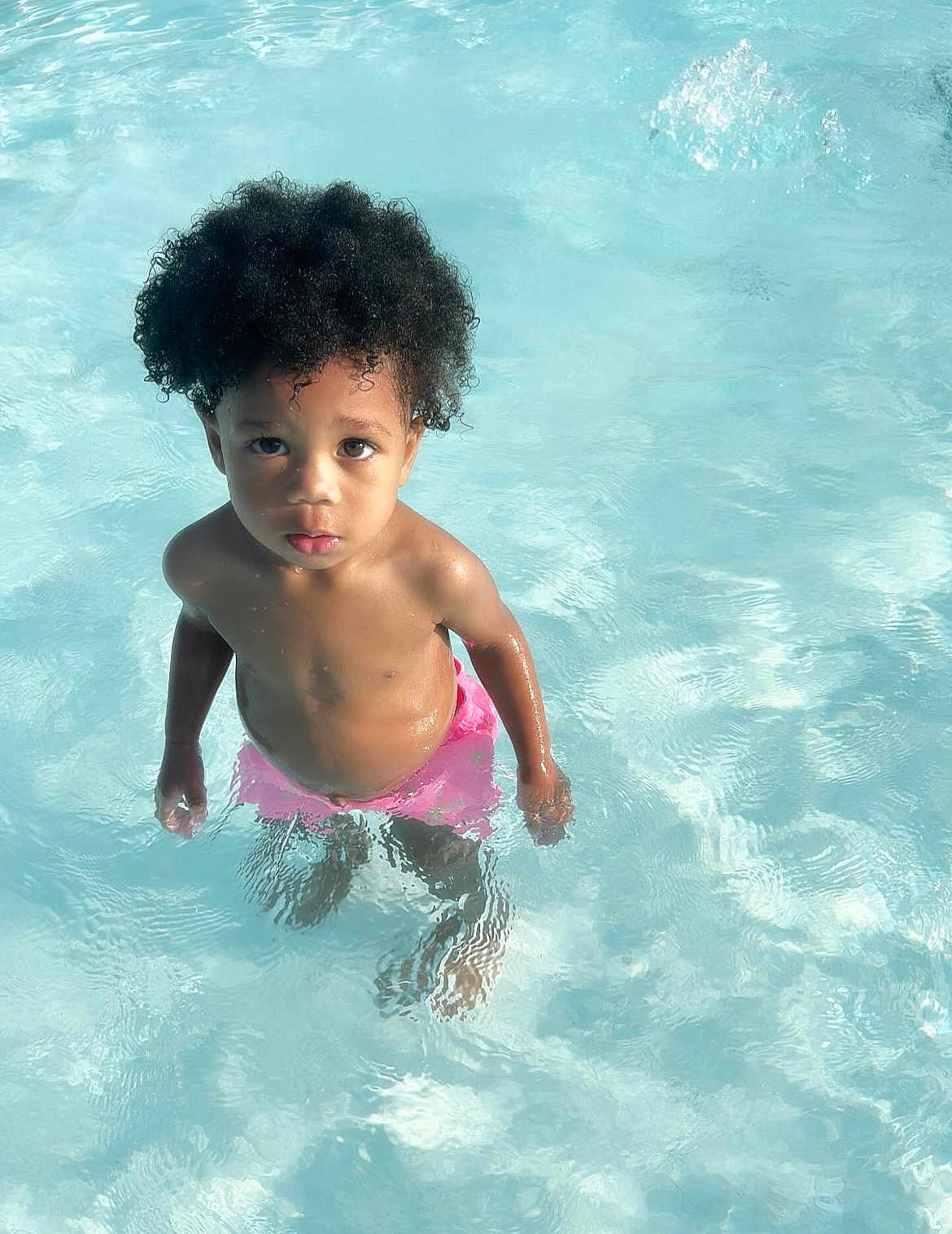 Kynd is registered to the contest to win money with this photo: child, toddler, water, pool, swimming, curly_hair, pink_shorts, summer, outdoor, wet, skin, standing, looking_up, innocent, young, water_ripples, sunlight, playful, vacation, fun