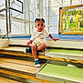 child, toddler, stairs, indoor, braided_hair, casual_clothing, blue_shoes, window, modern_architecture, stained_glass, wood, metal, handrail, curious, sitting, expression, light, daylight, young_child, person