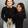 black_shirt, casual_clothing, cat, face_touch, friendship, gray_cat, happy, holding_cat, indoor, jeans, light_skin, long_hair, medium_skin, portrait, posing, short_hair, smiling, studio_background, tabby_cat, two_people