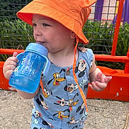 Maël participe au concours pour gagner de l'argent avec cette photo : toddler, child, hat, orange_hat, sippy_cup, blue_cup, mickey_mouse, cartoon_clothing, outdoor, fence, greenery, playground, smiling, cute, summer_clothes, baby, person, daylight, standing, casual