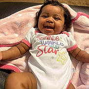 Kaliah joined the competition — help win amazing prizes! baby, blanket, child, closeup, clothing, curly_hair, cute, face, happy, indoors, infant, joyful, lying_down, onesie, person, pink, smiling, soft_texture, star, toddler