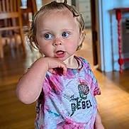 Penny is registered to the contest to win money with this photo: toddler, child, person, indoor, tie_dye_shirt, curly_hair, top_knot, blue_eyes, hand_near_mouth, wooden_floor, furniture, shirt, colorful, expression, portrait, standing, home, casual_clothing, young_child, cute