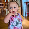 toddler, child, person, indoor, tie_dye_shirt, curly_hair, top_knot, blue_eyes, hand_near_mouth, wooden_floor, furniture, shirt, colorful, expression, portrait, standing, home, casual_clothing, young_child, cute