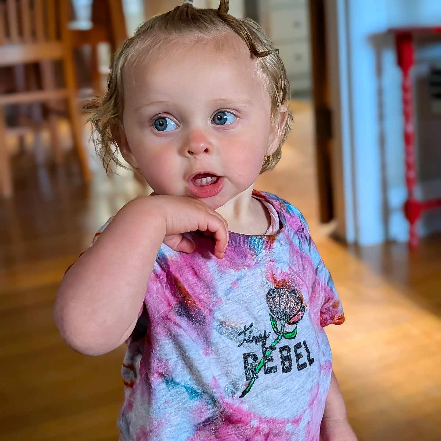 Penny is registered to the contest to win money with this photo: toddler, child, person, indoor, tie_dye_shirt, curly_hair, top_knot, blue_eyes, hand_near_mouth, wooden_floor, furniture, shirt, colorful, expression, portrait, standing, home, casual_clothing, young_child, cute