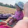 adult_arm, barefoot, beach, casual, child, outdoor, pink_clothing, playful, purple_towel, relaxed, sand, sippy_cup, summer, sunhat, sunny, tattoo, toddler, towel, trees, unicorn