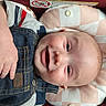 baby, smiling, denim_overalls, striped_shirt, car_seat, red_fabric, infant, happy, child, cute, portrait, clothing, face, skin, head, small_hand, button, text_warning_label, indoor, seat