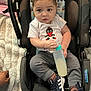 baby, bottle, car_seat, clothing, footwear, indoor, person, sitting, toy, toddler, white_shirt, gray_pants, sneakers, hand, blanket, fabric, face, young_child, milk, adidas