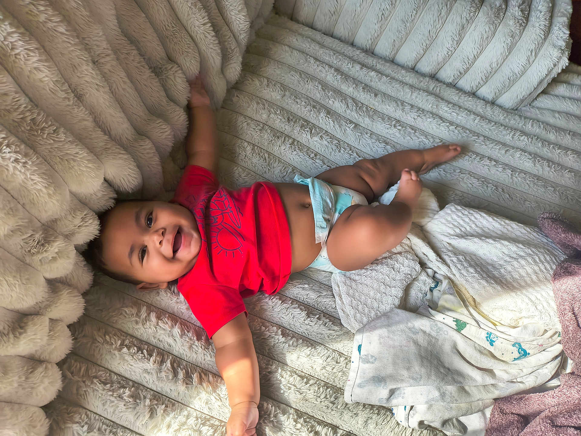 Kash is registered to the contest to win money with this photo: baby, infant, smiling, red_shirt, diaper, blanket, soft_texture, cozy, lying_down, happy, child, cute, plush, comfort, indoor, person, toddler, reaching, skin, head