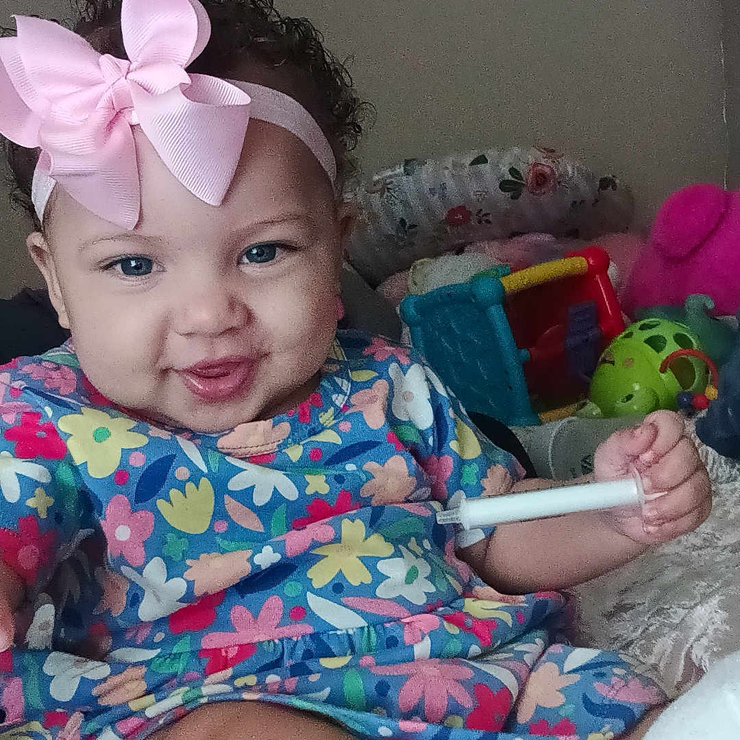 Jarianna is registered to the contest to win money with this photo: baby, child, smiling, pink_bow, curly_hair, colorful_dress, toys, indoor, cushion, playtime, happy, person, face, hand, foot, sock, flower_pattern, headband, cute, seated