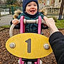 Marin participe au concours pour gagner de l'argent avec cette photo : child, toddler, smile, playground, play_equipment, beanie, winter_coat, hands, adult_hand, number_one, rocker, wood_chips, outdoor, fence, house, happy, cheeks, teeth, park, cold_weather