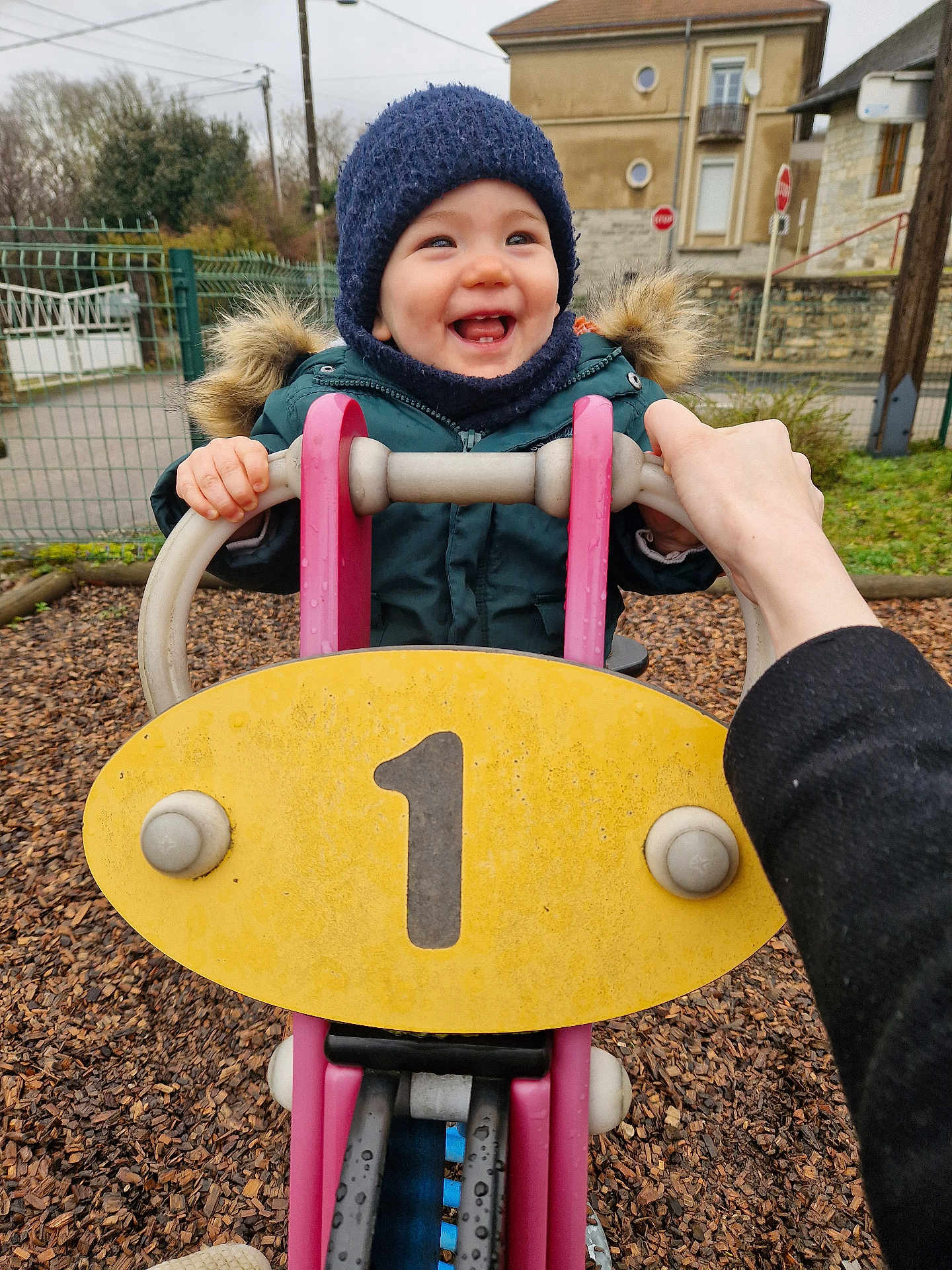Marin participe au concours pour gagner de l'argent avec cette photo : child, toddler, smile, playground, play_equipment, beanie, winter_coat, hands, adult_hand, number_one, rocker, wood_chips, outdoor, fence, house, happy, cheeks, teeth, park, cold_weather