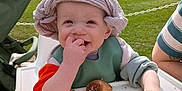 Erik is registered to the contest to win money with this photo: baby, child, toddler, highchair, hat, bib, bread_roll, eating, smiling, crumbs, hand, outdoor, park, grass, stroller, picnic, sunlight, portrait, headwear, happy