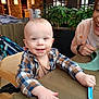 Erik joined the competition — help win amazing prizes! baby, woman, smile, high_chair, stroller, indoor, restaurant, table, spoon, plaid_shirt, plants, joy, parent, hand, wooden_table, face, laughing, casual_clothing, feeding, cozy